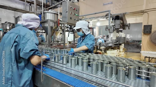 Several metal cans are lined up on a conveyor in a production facility. Workers in the background monitor the machinery's operation, ensuring a smooth flow of the manufacturing process.