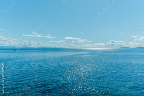 Fotografie Sparkling blue sea under a bright sky with distant mountains and a small sailboa
