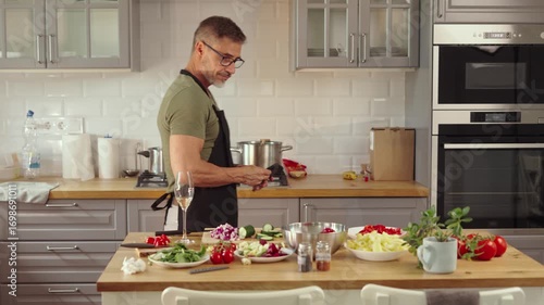 Mid adult man preparing for cooking in modern kitchen at home, wearing casual cloth and apron. Male chef following food recipe, cleaning and slicing vegetables, drinking champagne.