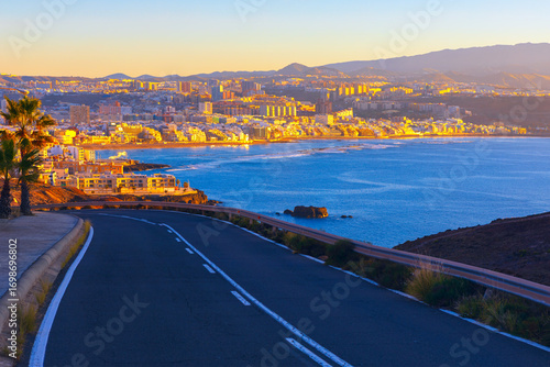 Sunset view of Las Palmas city in Gran Canaria with golden light reflecting on buildings along the coast. Winding road leads toward the Atlantic Ocean, framed by mountains in background