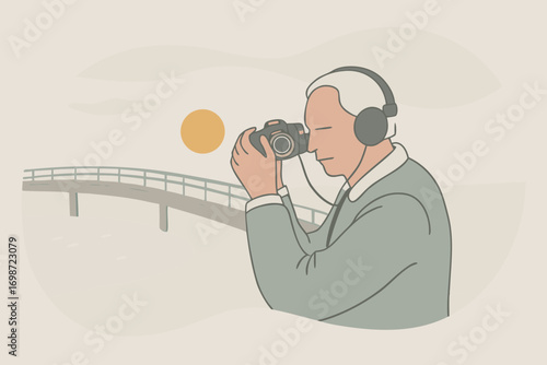 Elderly man with headphones and camera around neck, standing on bridge, photographing sunset over river