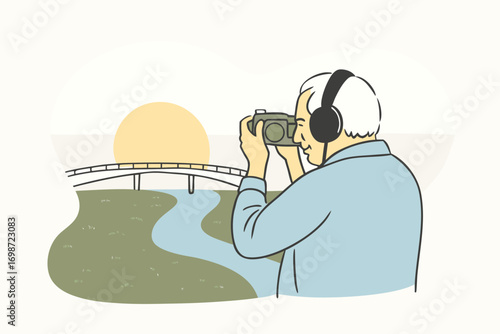 Elderly man with headphones and camera around neck, standing on bridge, photographing sunset over river.