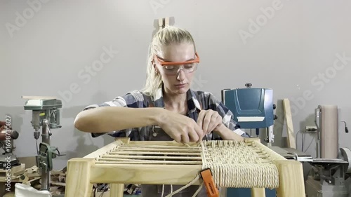 Crafting with Care: A focused craftswoman with safety glasses meticulously weaves a cord seat onto a wooden stool, surrounded by woodworking tools in a workshop.