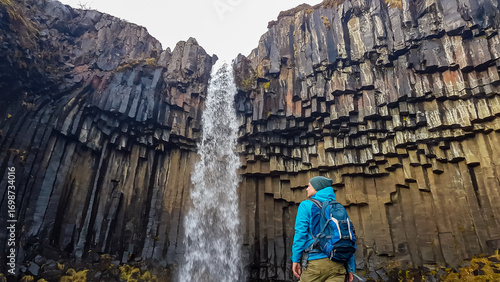 A male hiker with a backpack stands in awe, looking up at the majestic Svartifoss waterfall and the incredible surrounding wall of hexagonal basalt columns in Skaftafell National Park, Iceland.