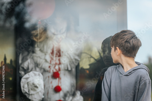 A young boy in a gray hoodie stares at a menacing clown figure with bloodstained costume and red balloons inside a glass display, creating a chilling horror-themed scene.