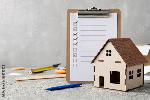A wooden house model sits beside a clipboard with a home inspection checklist and various essential tools
