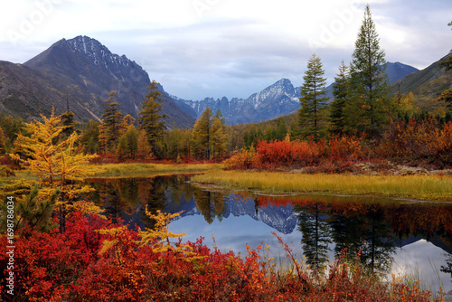 Autumn nature of Magadan, Kolyma. Colorful Taiga. Snowy mountains are reflected in the serene surface of the lake. A remote, hard-to-reach place for hiking, backpacking