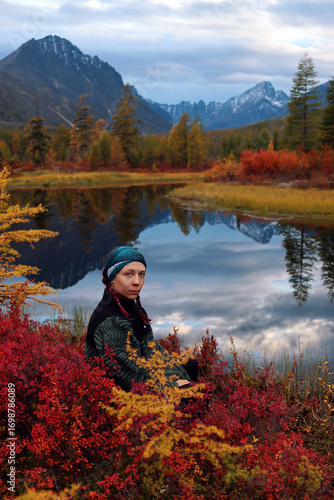 A young female hiker enjoys the views of the autumn taiga. A traveler is relaxing near a lake that displays snow-capped mountains. A forest painted with the bright colors of the changing season