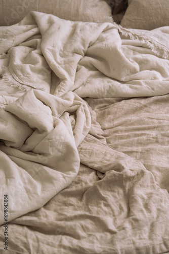 Double bed with crumpled oatmeal linen bedding and pillows, soft sunlight shadows creating a cozy aesthetic morning atmosphere.	