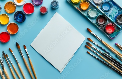 Watercolor Paints and Brushes Surrounding Blank White Paper on Blue Background