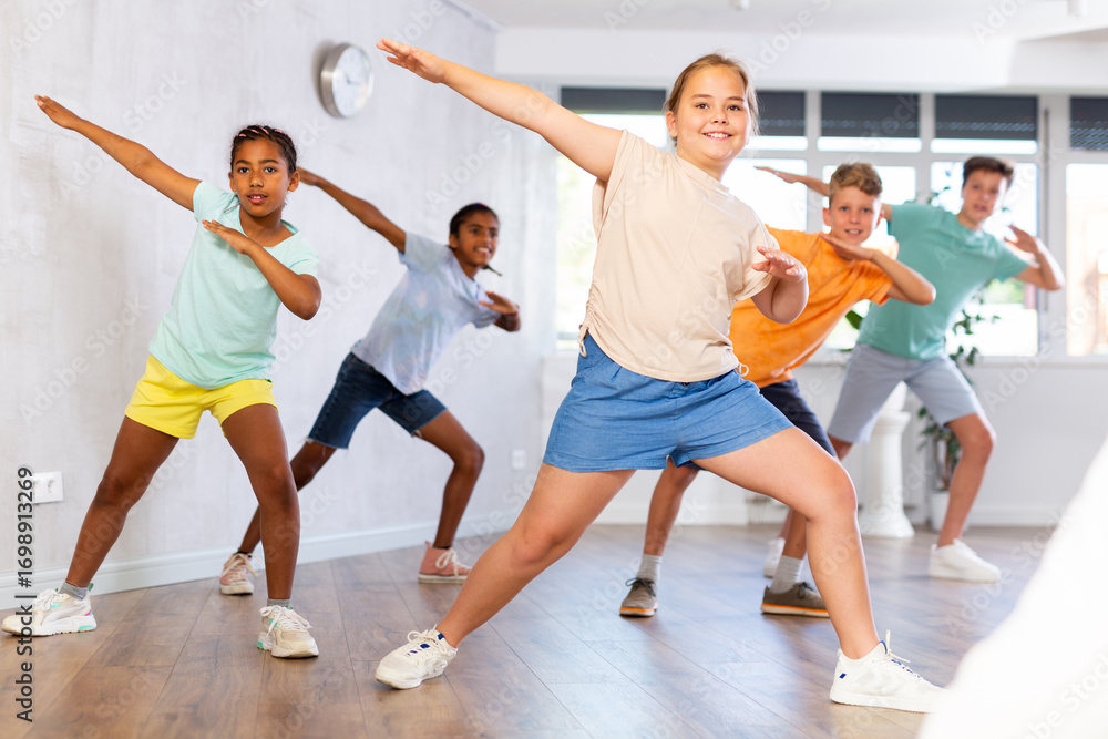 Fototapeta premium Smiling cheerful tween girl practicing modern dances with group of children in studio, doing dabbing move..