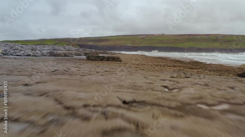 Low drone tracking shot along the wild Doolin coastline, with the roaring Atlantic crashing in and the Cliffs of Moher rising dramatically in the background.