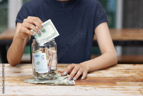 A person places dollar bills into a glass jar filled with cash on a wooden table, representing saving money, financial planning, and building future security through budgeting and smart spending.