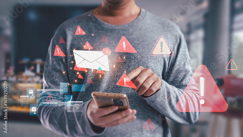 A man holds a smartphone while receiving a spam email notification. The screen displays red warning icons and system hacked alerts, illustrating a cybersecurity phishing scam concept.