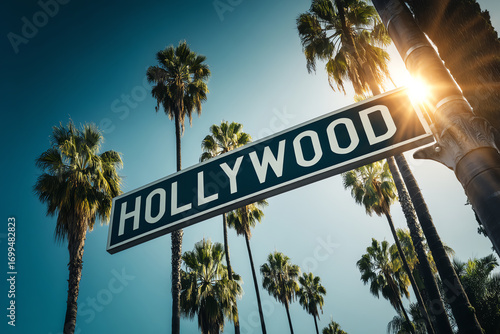 Hollywood street sign against a clear blue sky with tall palm trees and sunlight shining through, representing the iconic Los Angeles entertainment district