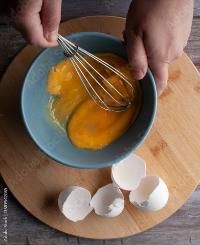 Female hands beating eggs inside a blue bowl on a round wooden cutting board, with the eggshells on one side, top view lay flat