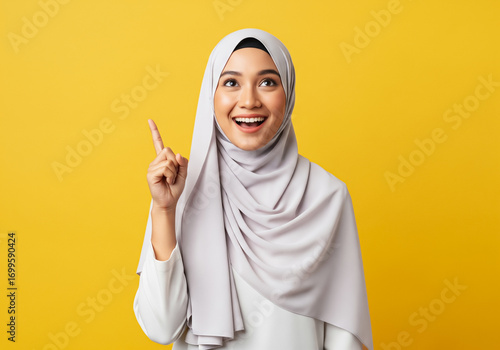 Excited beautiful Asian girl wearing hijab having creative idea, pointing finger up at copy space isolated on yellow background