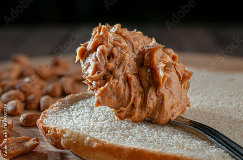 Canvas Print A spoonful of peanut butter on a slice of white bread on a wooden cutting board with peanuts out of focus in the background