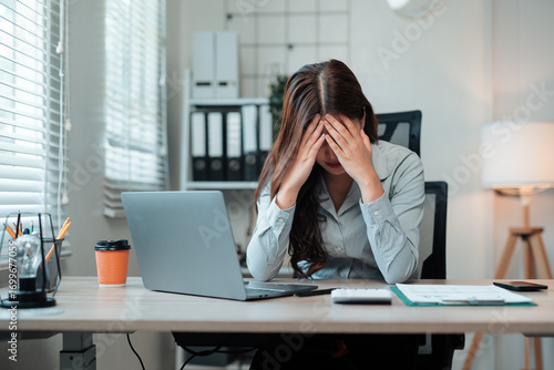 Young Asian businesswoman is overwhelmed by work and holds her head in her hands, sitting at her desk in the office