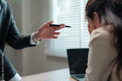 Business woman covering face with hands while manager is scolding her for bad work results pointing with pen sitting at office desk