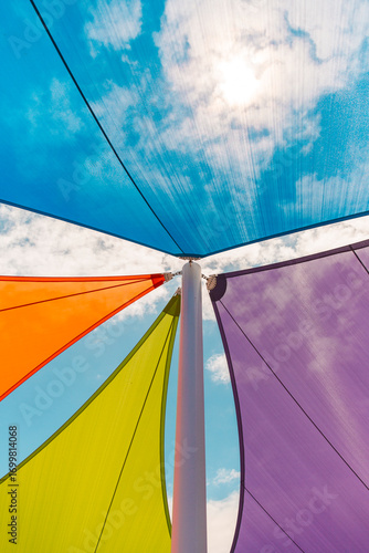 Colorful shade sails stretched above pole viewed from below, vibrant fabric triangles in blue, orange, green, and purple against bright sky
