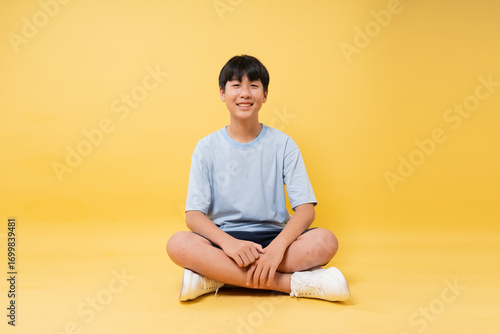 Smiling teen sits cross-legged on a bright yellow background wearing casual clothing and sneakers