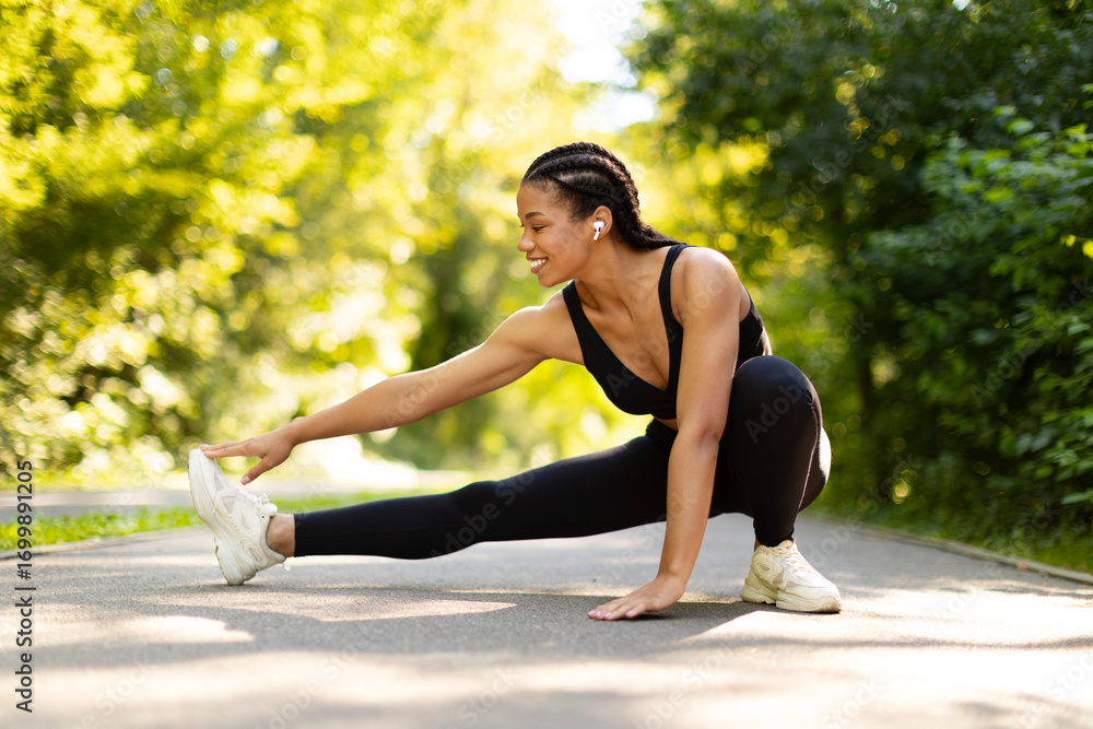 Fototapeta premium Happy African American woman stretching outdoors, smiling during fitness routine. Healthy lifestyle, energy, flexibility, motivation, and outdoor workout