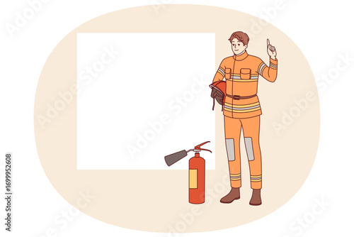 Man firefighter stands near fire extinguisher and copy of space for safety advice. Young guy firefighter from 911 service recommends being careful with electrical equipment and flammable materials