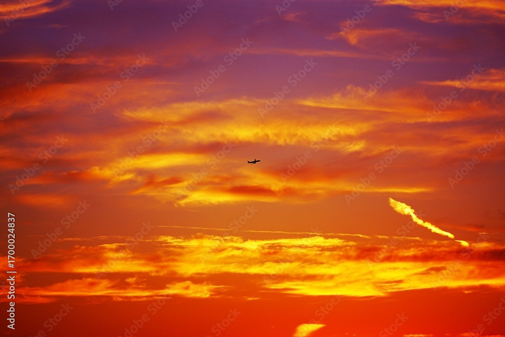 Naklejka premium Dramatic sky at sunset with airplane silhouette flying through colorful clouds. Fiery tones of orange, yellow and red blend creating breathtaking view. Concept of travel, freedom, wanderlust, aviation