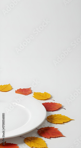 Elegant autumn dining concept featuring vibrant fall leaves surrounding a pristine white plate on white backdrop
