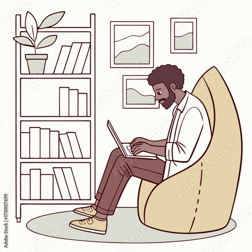 Bearded black man sitting in ergonomic chair, typing on laptop with bookshelf in background.