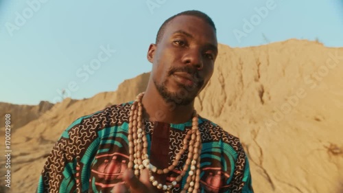 Young Black man wearing patterned ethnic outfit with wooden beads, playing djembe in semi-desert