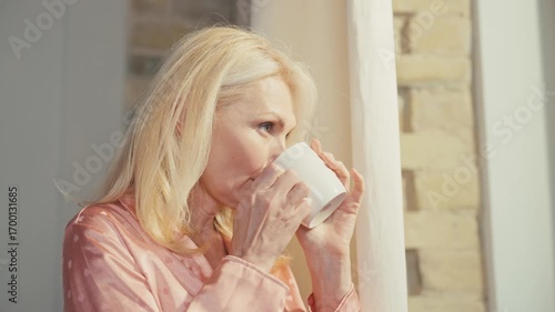Smiling Caucasian woman in peach pajama holding white cup and looking at camera. Standing near window with cheerful face. Enjoying hot coffee or tea while greeting new day with happiness.