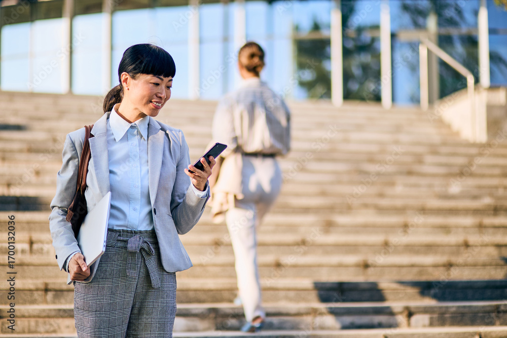 Fototapeta premium Asian Businesswoman Viewing Smartphone While Carrying Laptop on City Stairs
