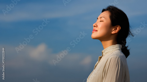 woman peacefully closing her eyes in calm pose against clear blue sky with fluffy clouds