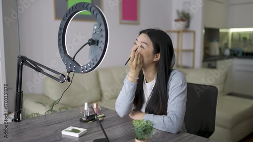 Young Thai woman recording a beauty video vlog at home. She talks to the camera with confidence while applying makeup and sharing skincare tips.