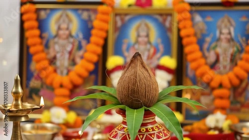 Image is a vibrant and colorful photograph depicting a traditional Indian religious setup. In the foreground, there is a kalash, a sacred pot, adorned with a coconut on top and surrounded by green man