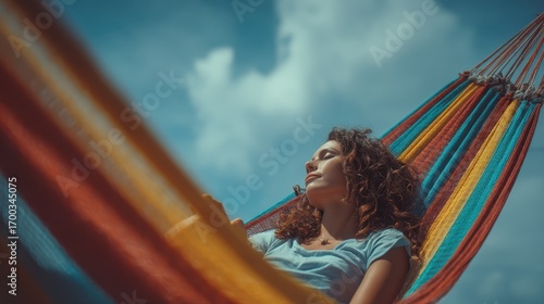 Young woman with curly hair relaxing with eyes closed in a colorful striped hammock on a sunny day under a clear blue sky