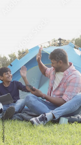 High-fiving Indian father and son sitting at campsite, with blue camping tent and soccer ball