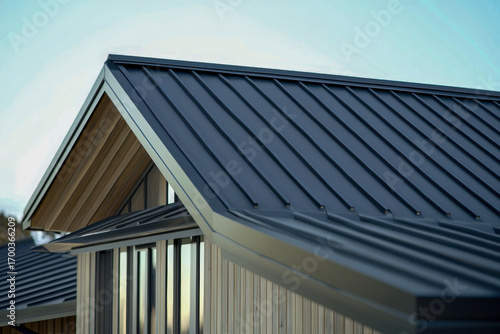 Modern house roof showing dark gray metal standing seam panels. Light wooden siding and eaves frame sharp architectural lines against clear blue sky. Exterior detail.