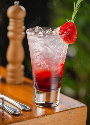 A refreshing strawberry cocktail served in a tall glass with ice, layered with red syrup, and garnished with a fresh strawberry slice and a rosemary sprig.