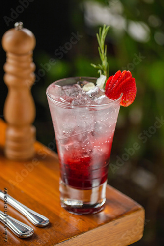 A refreshing strawberry cocktail served in a tall glass with ice, layered with red syrup, and garnished with a fresh strawberry slice and a rosemary sprig.
