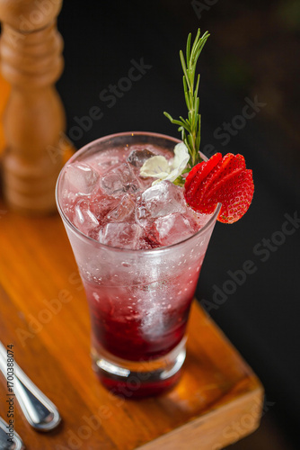 A refreshing strawberry cocktail served in a tall glass with ice, layered with red syrup, and garnished with a fresh strawberry slice and a rosemary sprig.