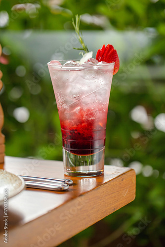 A refreshing strawberry cocktail served in a tall glass with ice, layered with red syrup, and garnished with a fresh strawberry slice and a rosemary sprig.