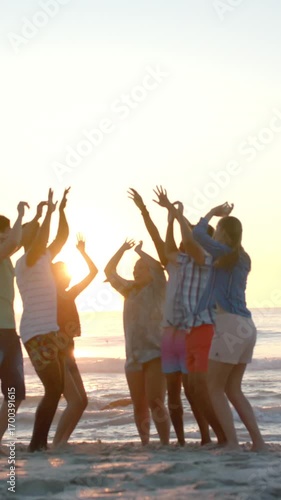 Vertical video: Diverse group of seven friends watching sunset lifting arms and bouncing on beach