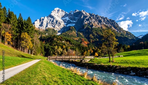 A serene mountain valley scene unfolds with a wooden bridge spanning a clear stream, vibrant autumn foliage, and snow-capped peaks rising above.