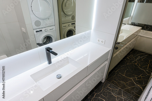 Modern bathroom featuring a sleek white vanity with a rectangular sink, black faucet, and a large mirror reflecting stacked laundry appliances. Dark marble flooring adds a luxurious touch