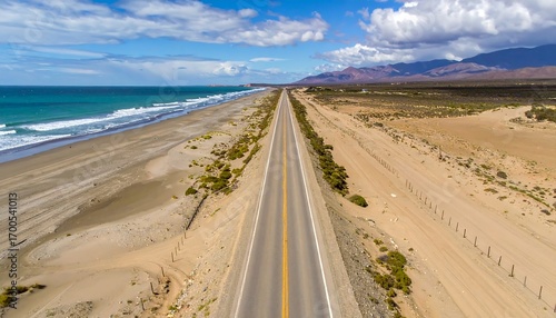 A paved highway stretches along a beach, bordered by dunes and a vast expanse of desert, under a vibrant, partly cloudy sky.