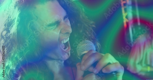 Singing curly-haired singer belting into microphone on club, with stage lights, truss and drum kit