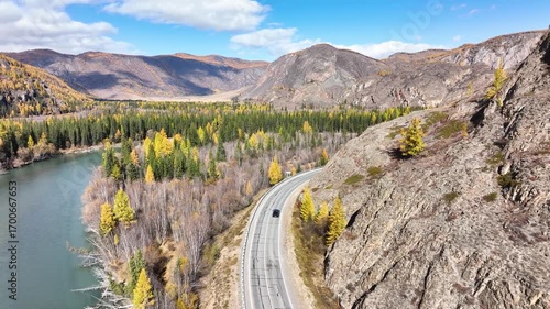A panoramic aerial view of a car driving on a winding road alongside a wide river, with surrounding mountains covered in vibrant golden autumn foliage under a clear blue sky